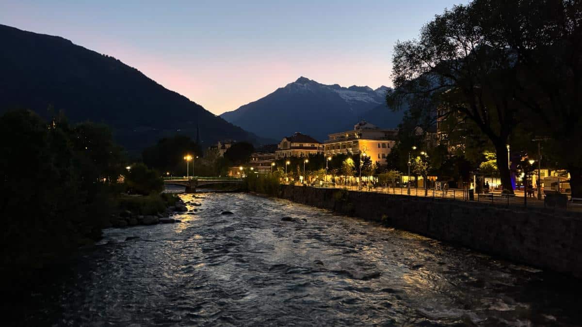 Sunset Over The Adige River Promenade In Merano, Italy, With Illuminated Buildings And Snowcapped Alps In The Background