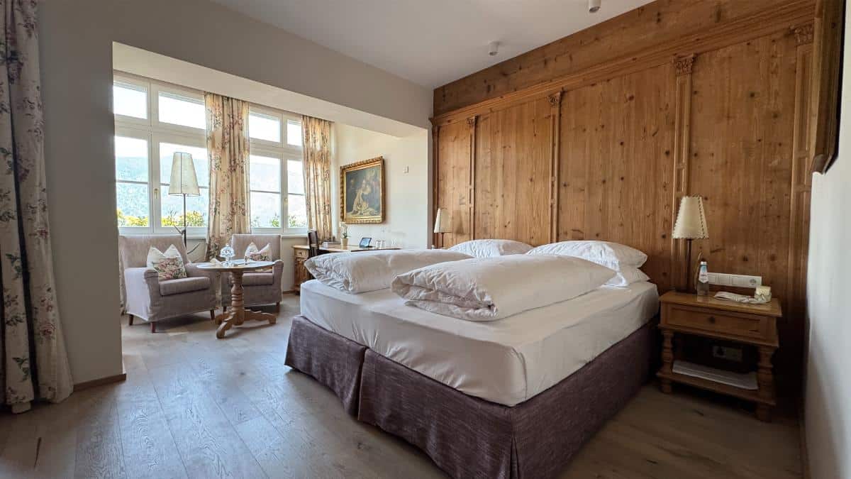 Junior Suite At Castel Fragsburg With Carved Wood Paneling, Plush King Bed, Floral Curtains, Sitting Area, And Mountain View Windows In Merano