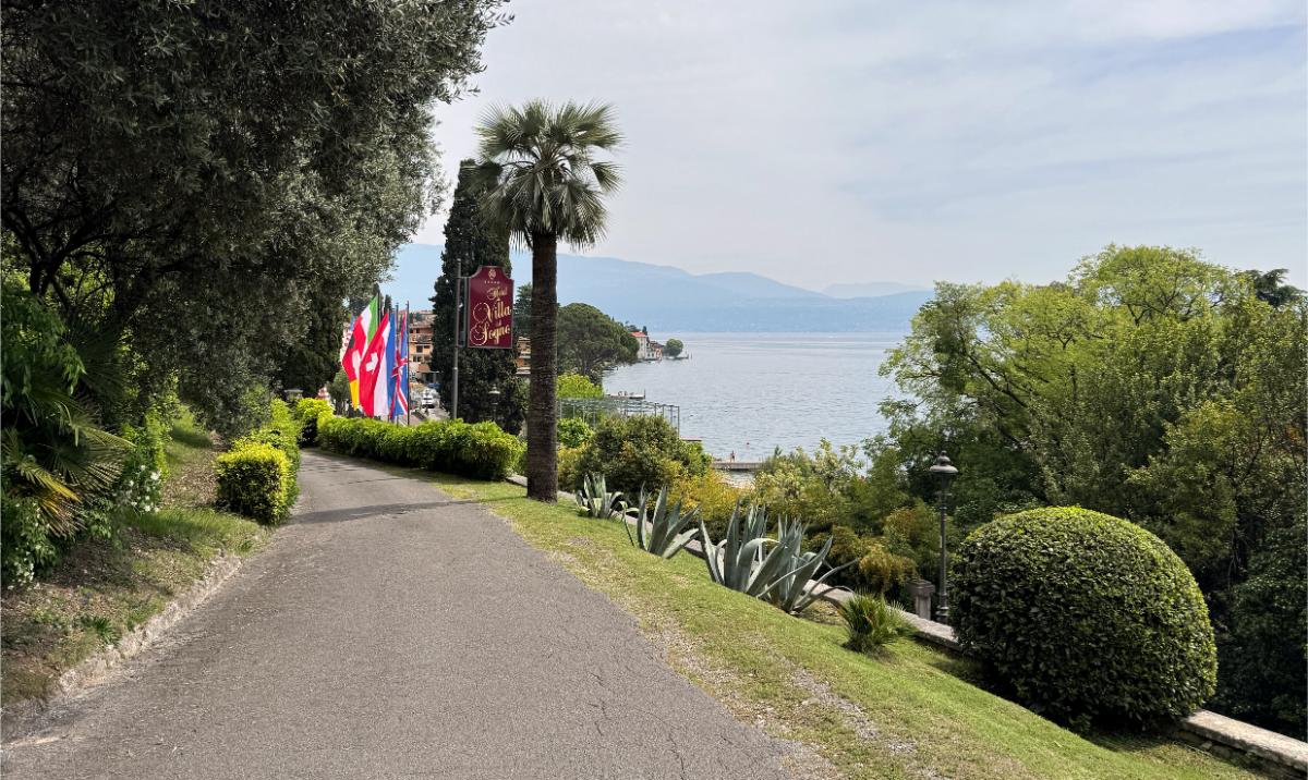 Tree-Lined Driveway Leading To Villa Del Sogno Hotel With The Property Sign, International Flags, And Lake Garda Visible In The Background